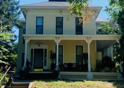 Two-story yellow house with white trim and a covered front porch, surrounded by greenery on a sunny day.