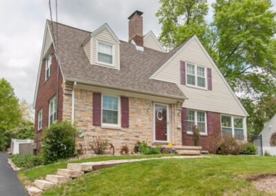 A charming two-story house with brick and stone accents, featuring a landscaped front yard and a cloudy sky overhead.