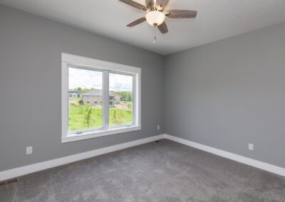 An empty room with gray walls, carpeted flooring, a ceiling fan, and a window with a view of a residential area.