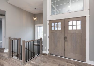 A modern home interior featuring wooden double doors with glass panels, a staircase with dark balusters and wooden handrail, and gray walls illuminated by natural light.